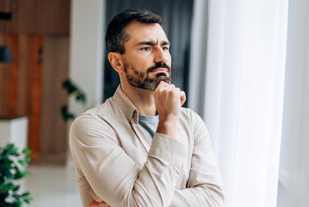 mężczyzna w wieku 30 plus z ciemnymi siwiejącymi włosami z zarostem w jasnej koszuli stoi przy oknie zamyślony