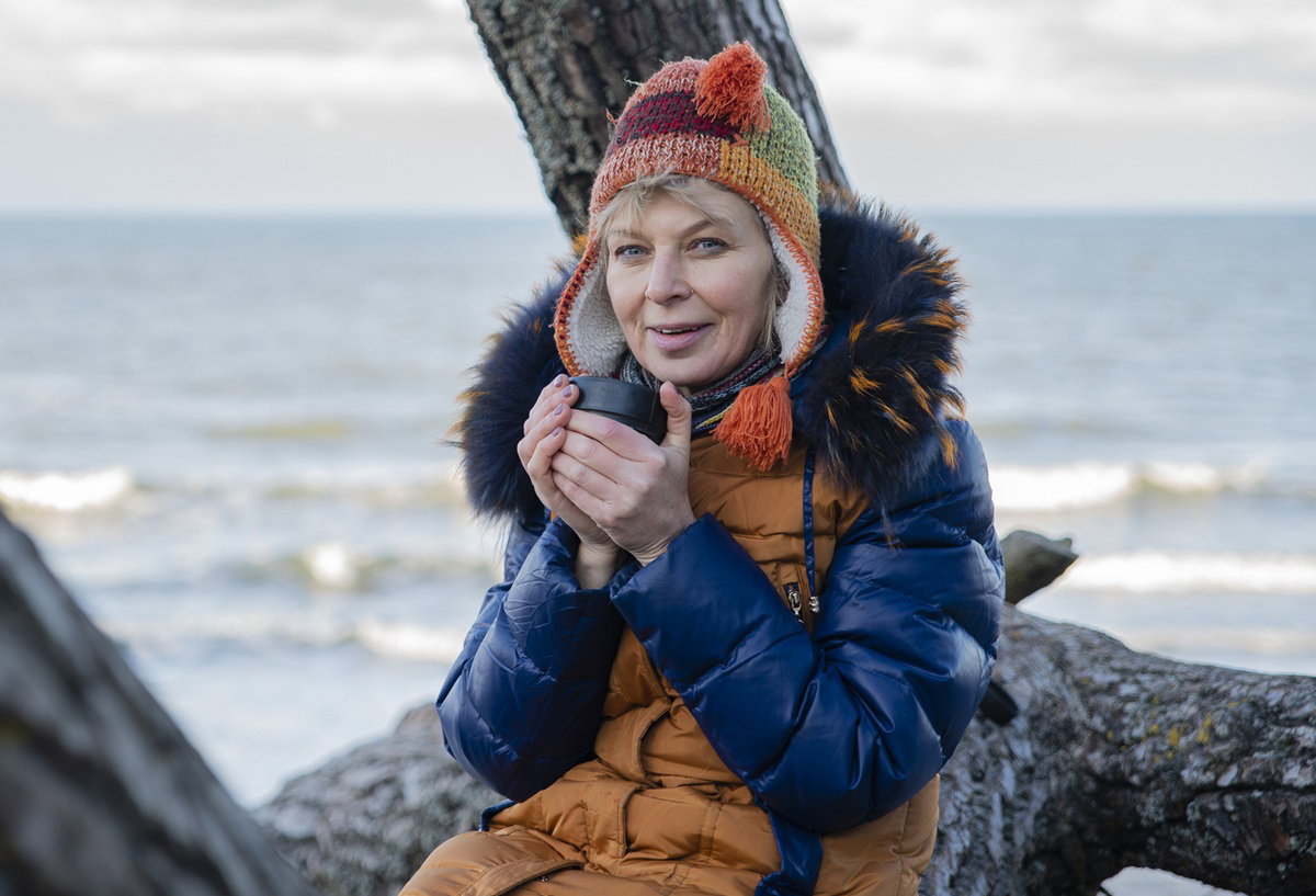 kobieta w wieku 50 plus w kolorowej czapce siedzi na tle morza i się uśmiecha