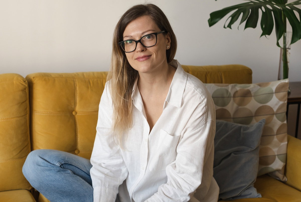 kobieta w wieku 35 plus w białej koszuli siedzi na kanapie i lekko się uśmiecha