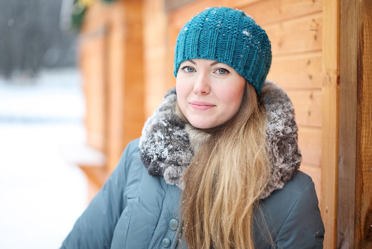 kobieta w wieku 30 plus w czapce i z długimi włosami stoi na tle drewnianego domu i delikatnie się uśmiecha