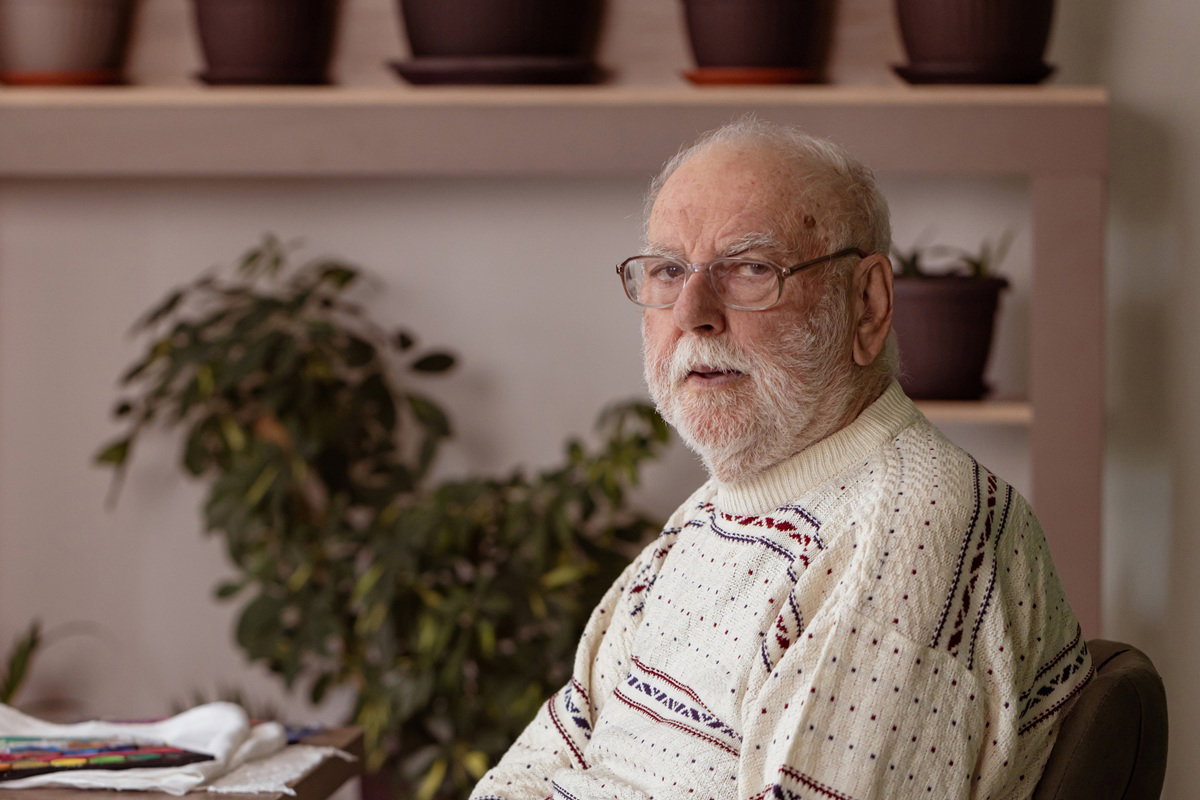 mężczyzna w wieku 70 plus w jasnym swetrze i w okularach siedzi smutny przy stole na tle roślin