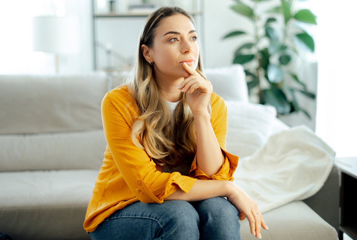 kobieta w wieku 35 plus w żółtej bluzce siedzi na kanapie zamyślona