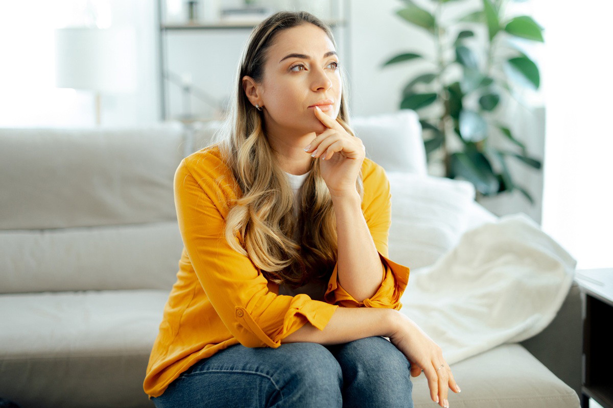 kobieta w wieku 35 plus w żółtej bluzce siedzi na kanapie zamyślona