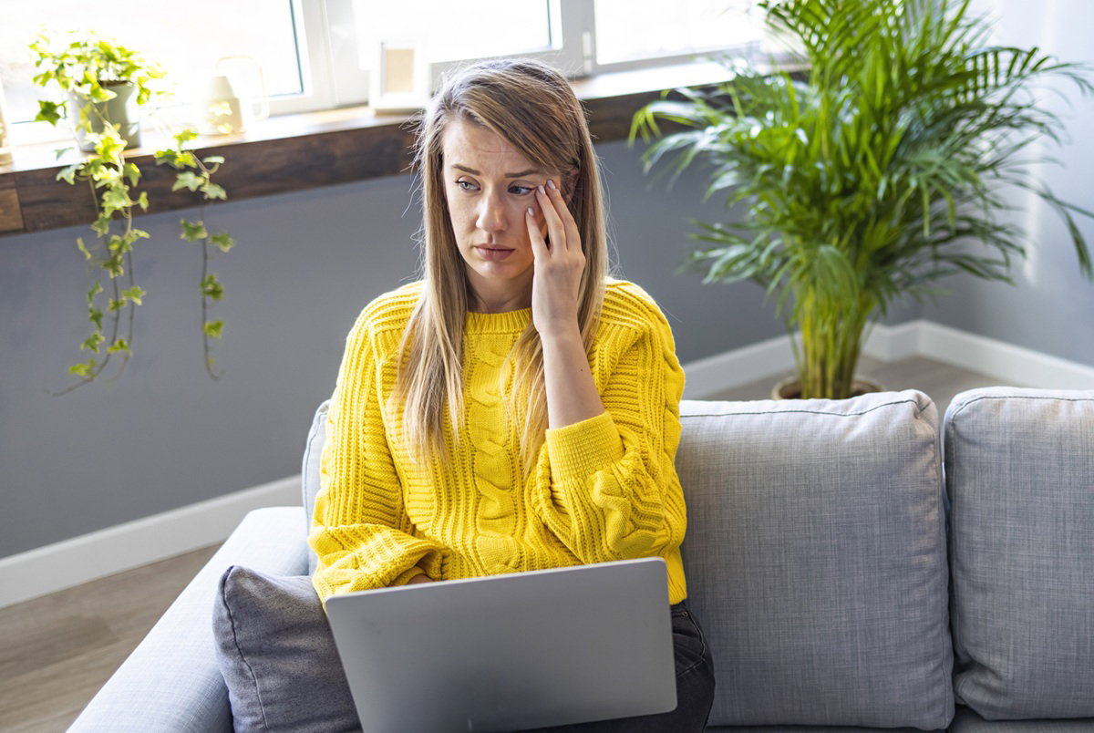 kobieta w wieku 30 plus w żółtym sweterku blondynka siedzi na kanapie z laptopem na kolanach załamana