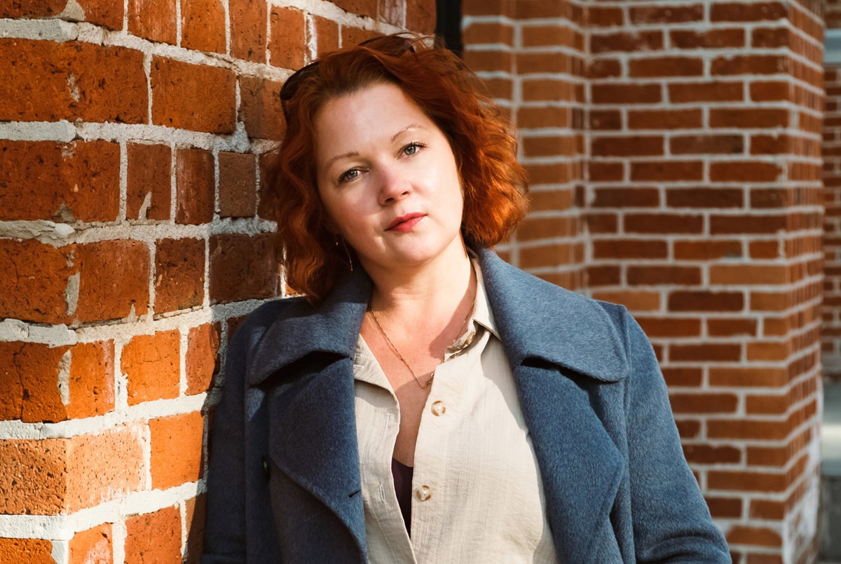 kobieta w wieku około 40 lat z rudymi włosami stoi na tle budynku z cegieł zamyślona i smutna