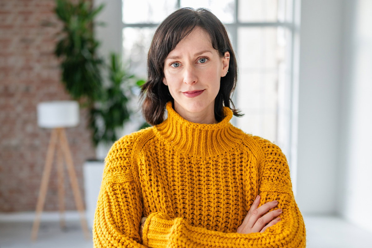 kobieta w wieku 40 plus w żółtym swetrze stoi na tle okna ze skrzyżowanymi ramionami