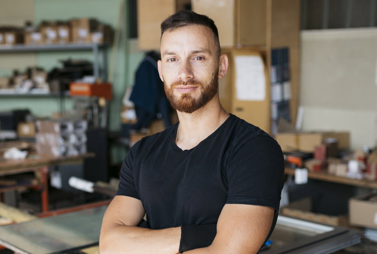 mężczyzna w wieku 30 plus z wąsami i brodą w czarnym t-shircie stoi na tle garażu z poważną miną