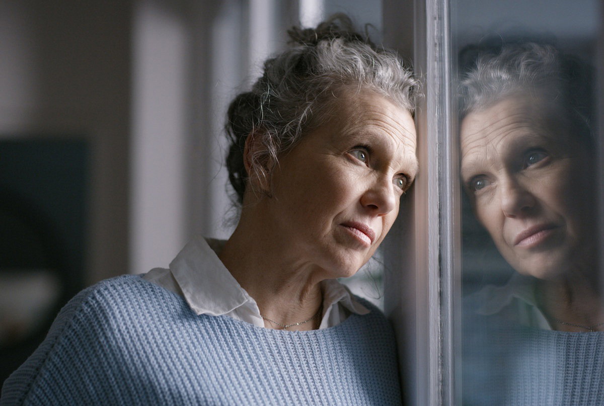 kobieta w wieku 60 plus siwa ze spiętymi włosami stoi zmartwiona przy oknie