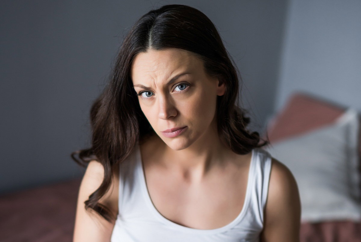 kobieta w wieku 30 plus z długimi włosami ze zmartwioną miną siedzi na łóżku