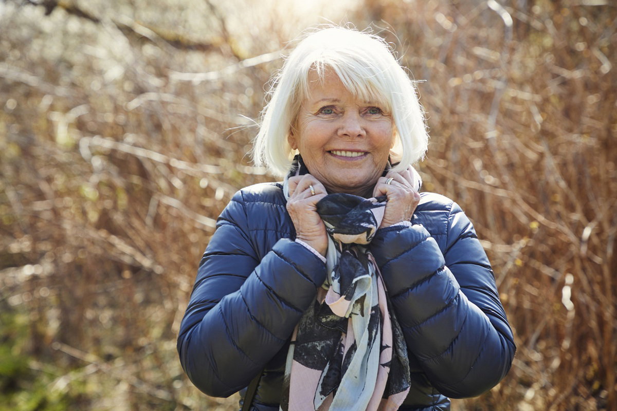 kobieta w wieku 70 plus z siwymi włosami stoi w parku i się uśmiecha zaskoczona