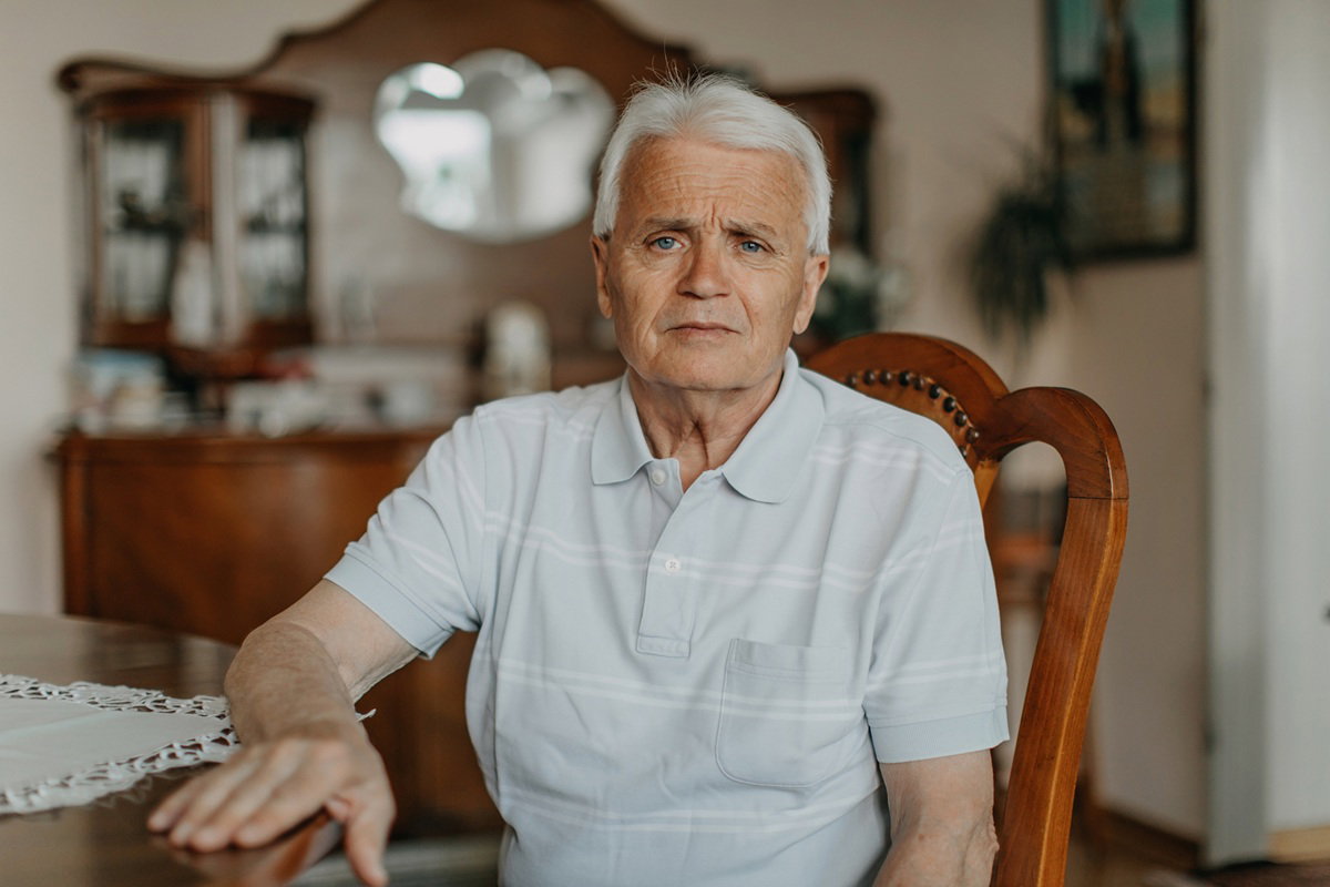 mężczyzna w wieku 65 plus w koszulce polo siedzi przy stole na krześle w salonie z poważną miną