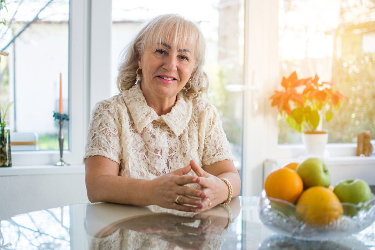 kobieta w wieku 60 plus w koronkowej bluzce siedzi przy stole uśmiechnięta