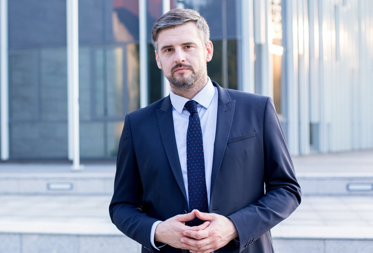 mężczyzna w wieku 35 plus w garniturze siwiejący stoi na tle szklanego biurowca z poważną miną