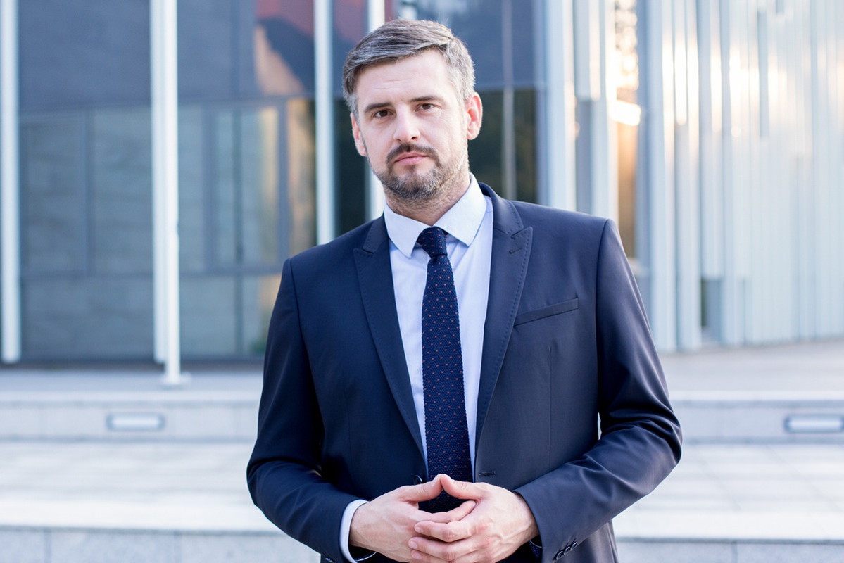 mężczyzna w wieku 35 plus w garniturze siwiejący stoi na tle szklanego biurowca z poważną miną