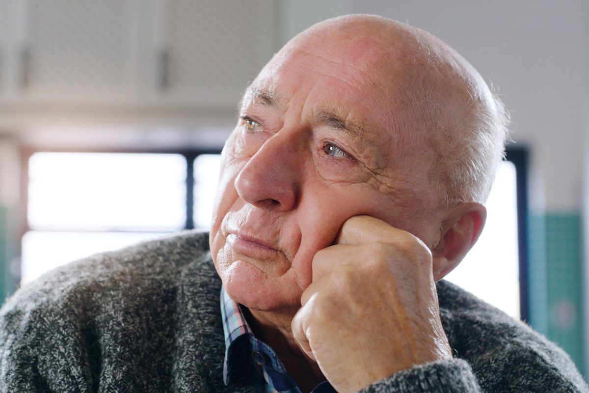 mężczyzna w wieku 65 plus siedzi na tle okna zmartwiony