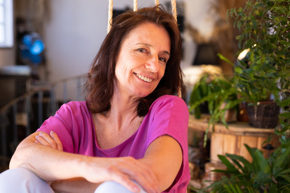 kobieta w wieku 45 plus w różowej bluzce siedzi i uśmiecha się szeroko na tle roślin