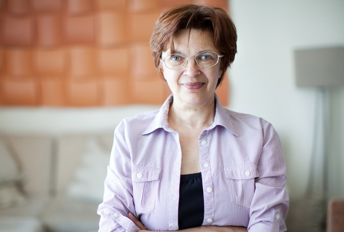 kobieta w wieku 50 plus w okularach stoi na tle salonu z niepewnym uśmiechem