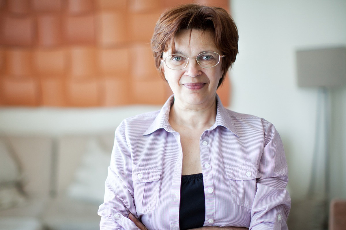 kobieta w wieku 50 plus w okularach stoi na tle salonu z niepewnym uśmiechem