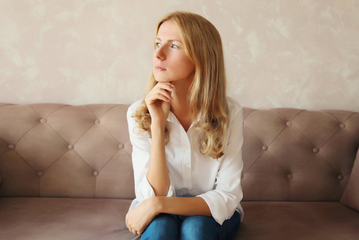 kobieta w wieku 30 plus blodnynka w białej koszuli siedzi na kanapie zamyślona