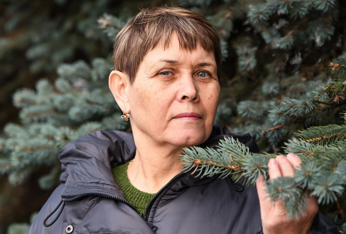 kobieta w wieku 55 plus w czarnej kurtce stoi na tle iglaków z poważną miną