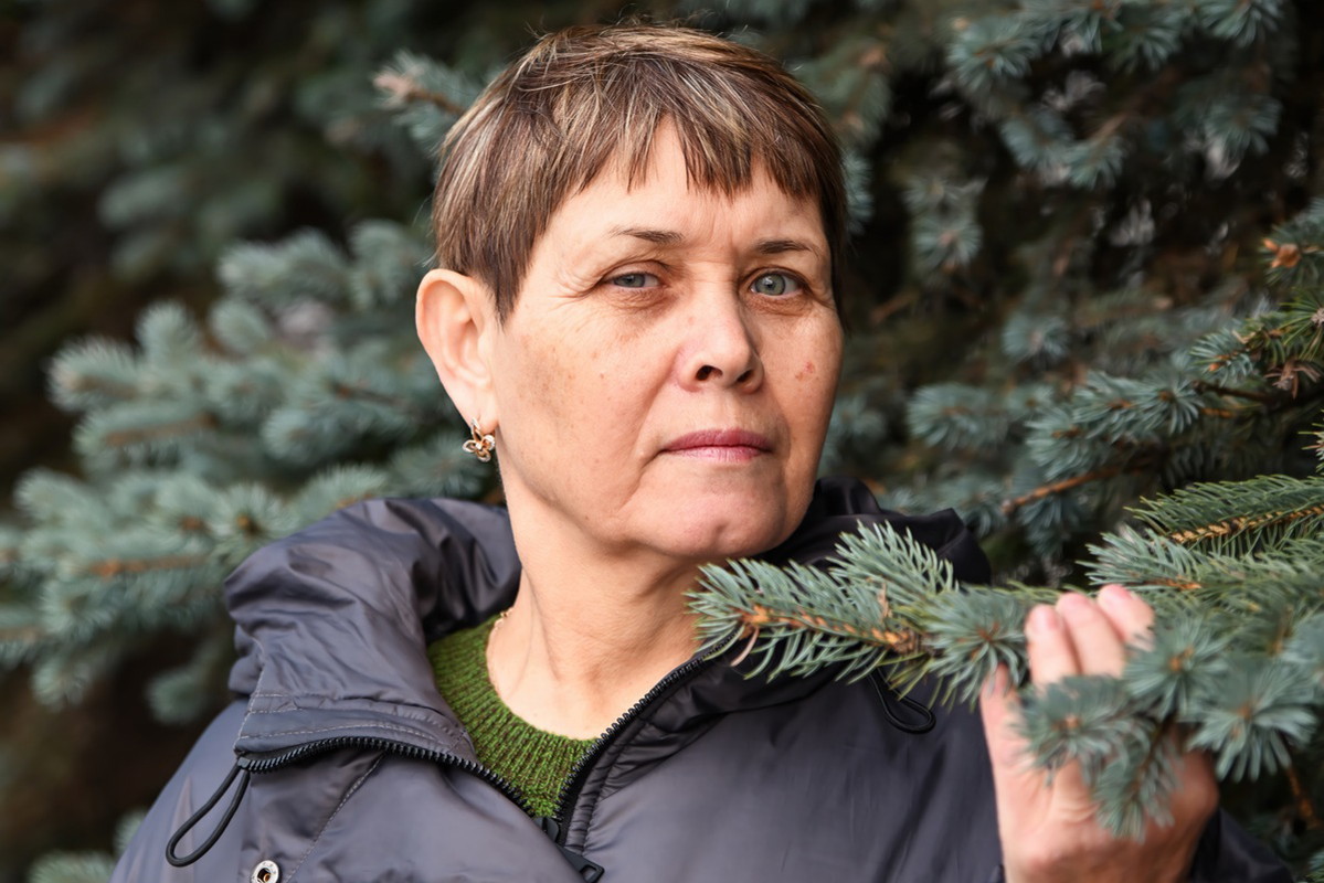 kobieta w wieku 55 plus w czarnej kurtce stoi na tle iglaków z poważną miną