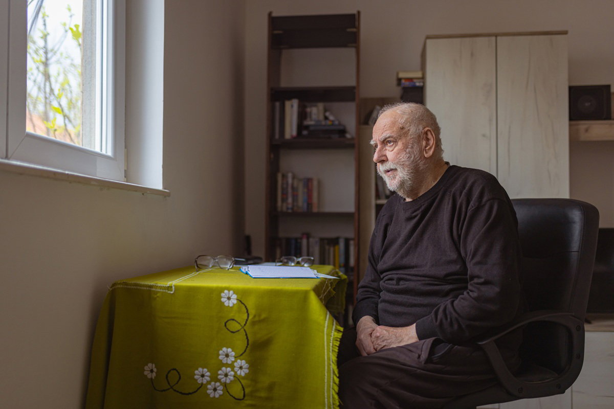 mężczyzna w wieku 75 plus w ciemnym ubraniu siedzi w pokoju i patrzy zamyślony w okno