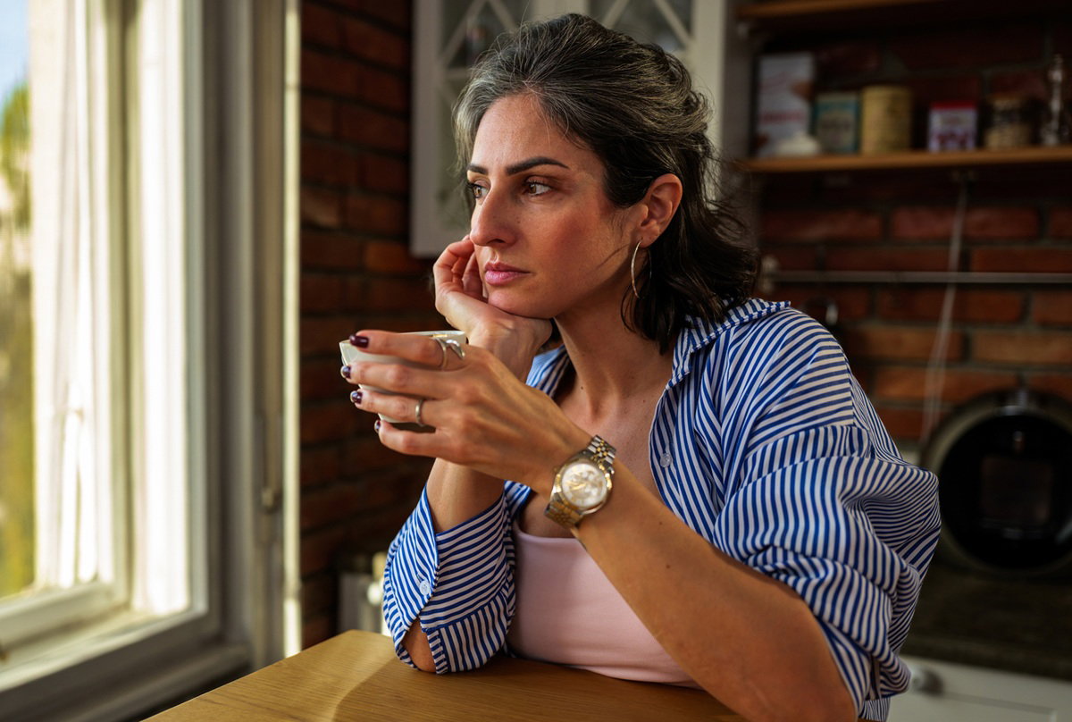 kobieta w wieku 40 plus w koszuli w paski siedzi przy kuchennym stole zamyślona i patrzy w okno