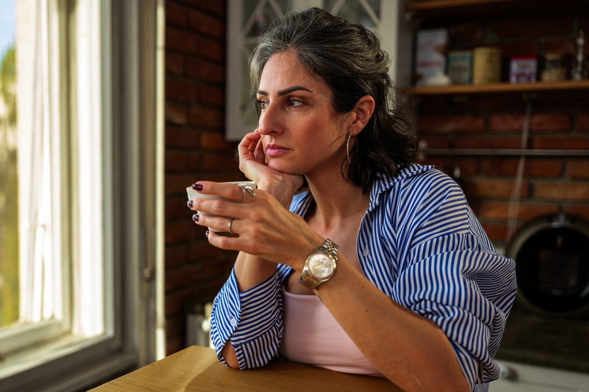 kobieta w wieku 40 plus w koszuli w paski siedzi przy kuchennym stole zamyślona i patrzy w okno