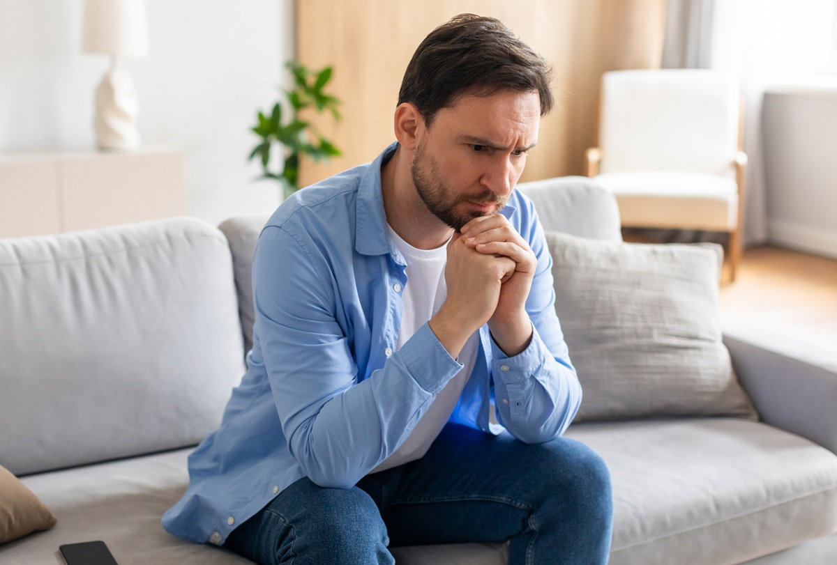 mężczyzna w wieku 35 plus w jasnej koszuli siedzi na kanapie zmartwiony