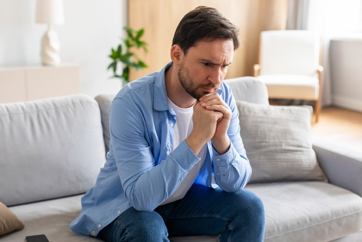 mężczyzna w wieku 35 plus w jasnej koszuli siedzi na kanapie zmartwiony