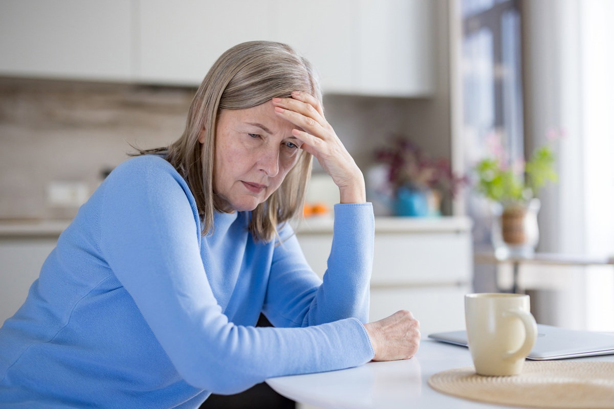 kobieta w wieku 60 plus w niebiskim sweterku siedzi przy stole w kuchni smutna