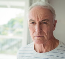 mężczyzna w wieku 60 plus siwy w szarym t-shircie w prążki stoi na tle okna z poważną miną