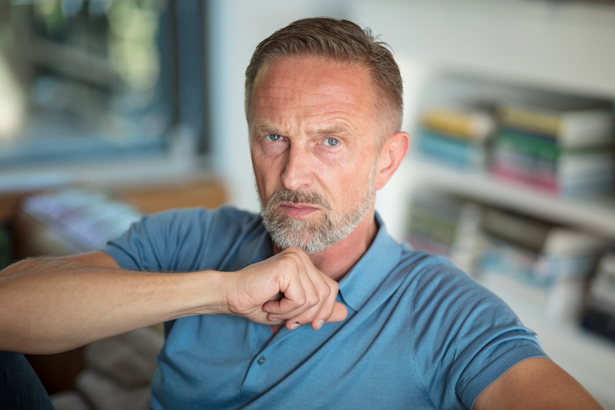 mężczyzna w wieku 45 plus siwiejący z wąsami i bródką siedzi zamyślony w salonie