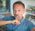 mężczyzna w wieku 45 plus siwiejący z wąsami i bródką siedzi zamyślony w salonie