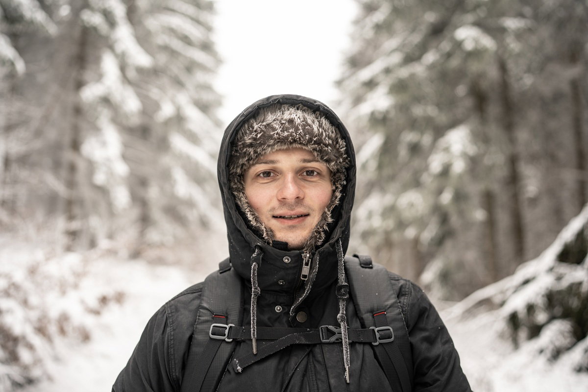 mężczyzna w wieku 30 plus w kapturze i uszance stoi lekko uśmiechnięty na tle zimowego pejzażu