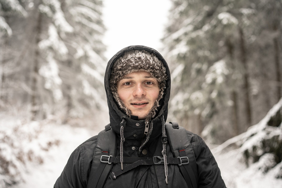 mężczyzna w wieku 30 plus w kapturze i uszance stoi lekko uśmiechnięty na tle zimowego pejzażu