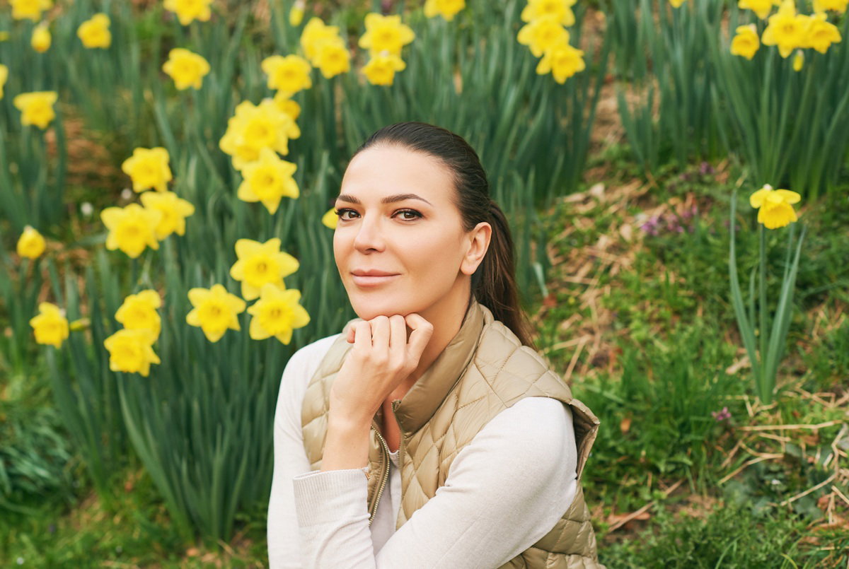 kobieta w wieku 30 plus siedzi na tle żonkili zamyślona z lekkim uśmieszkiem