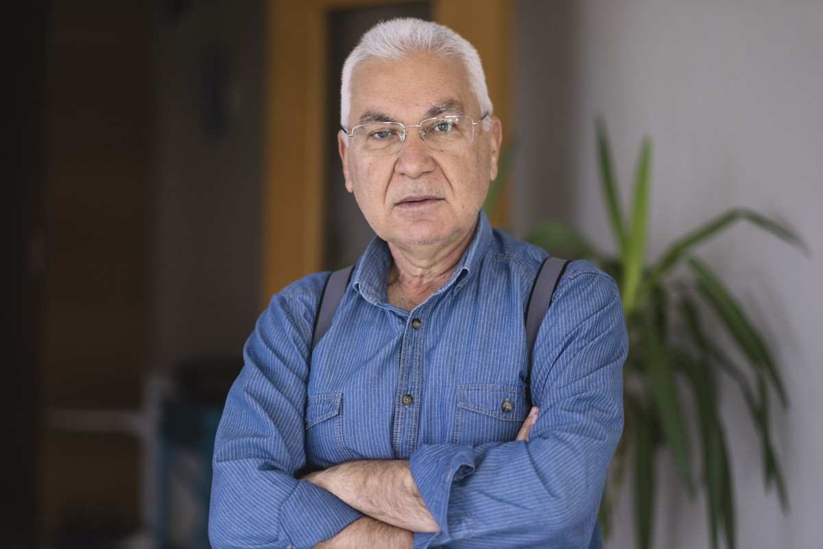 mężczyzna w wieku 65 plus siwy w okularach w niebieskiej koszuli stoi poważny na tle pokoju