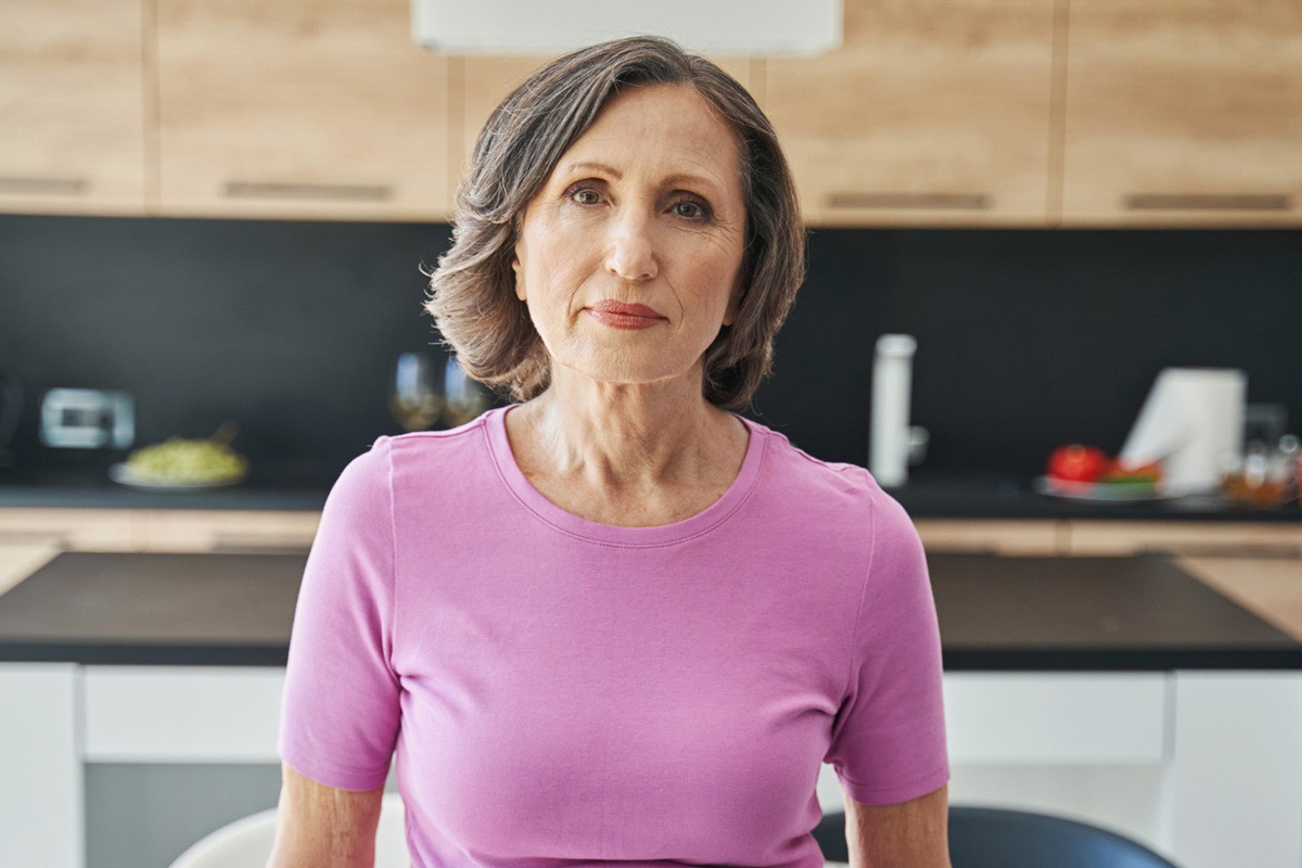 kobieta w wieku 60 plus w różowym t-shircie stoi na tle kuchni z poważną miną