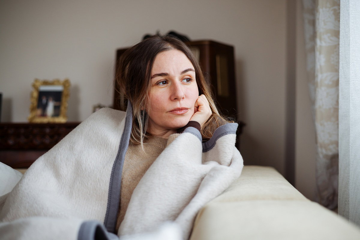 kobieta w wieku 35 plus siedzi na kanapie w kocu zamyślona