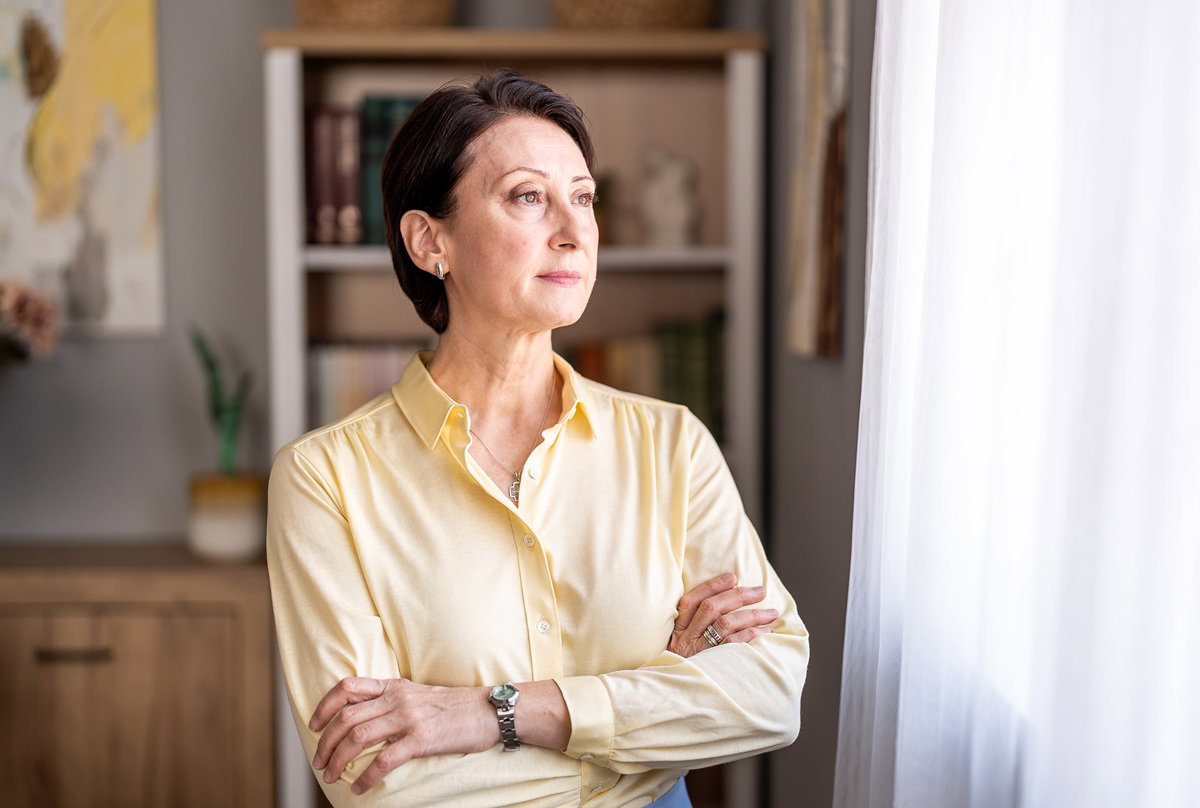 kobieta w wieku 50 plus w żółtej koszuli patrzy zamyślona w okno