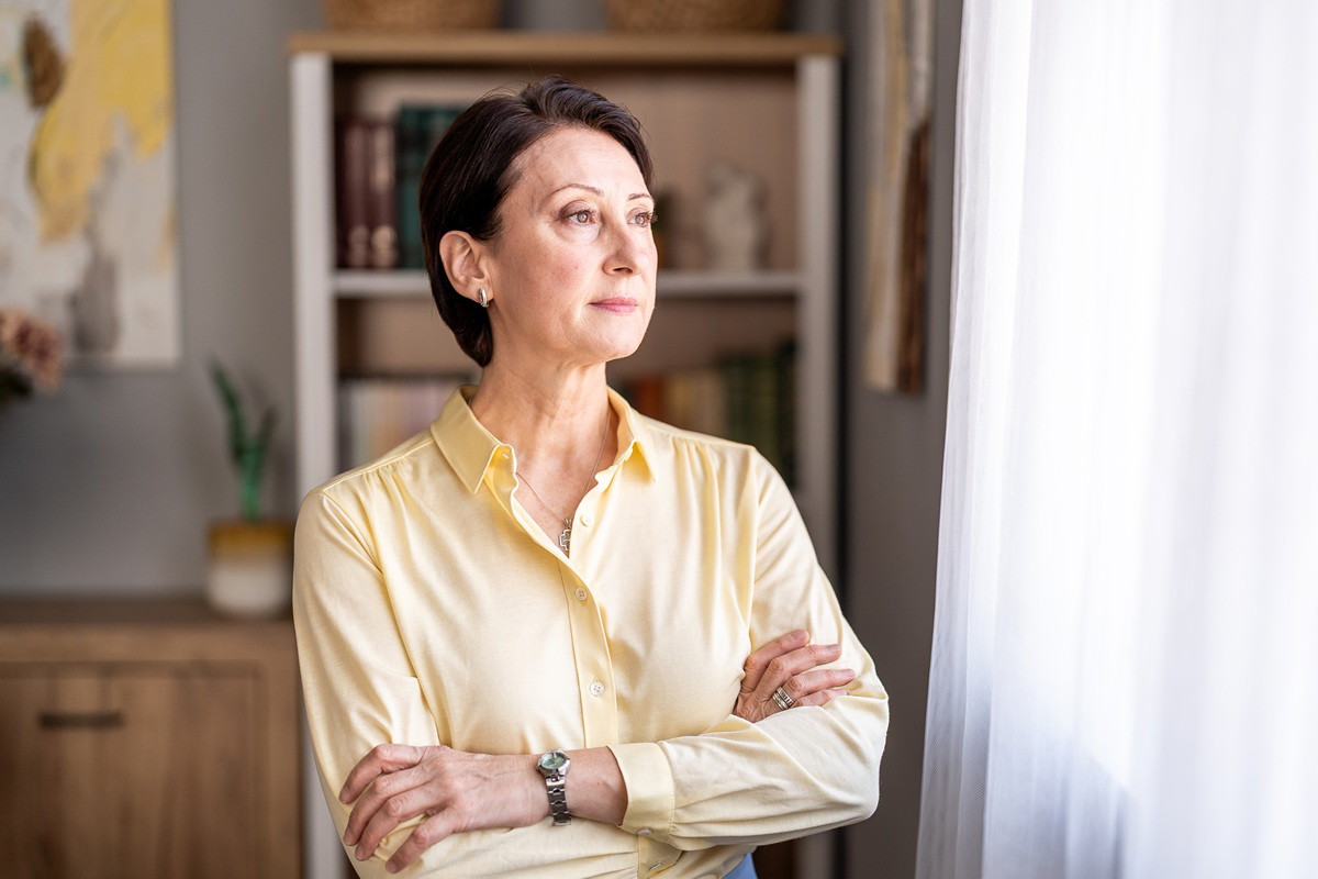 kobieta w wieku 50 plus w żółtej koszuli patrzy zamyślona w okno