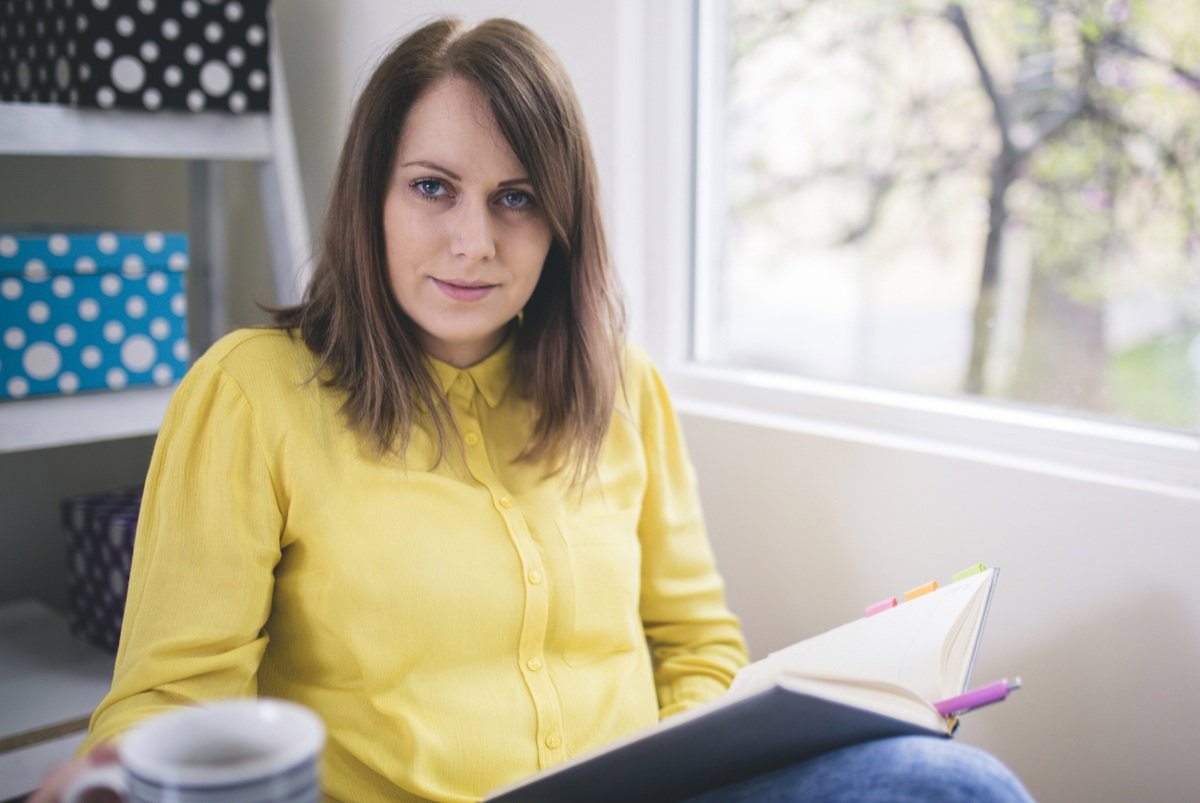 kobieta w wieku 40 plus w żółtej koszuli siedzi przy oknie z książką i herbatą