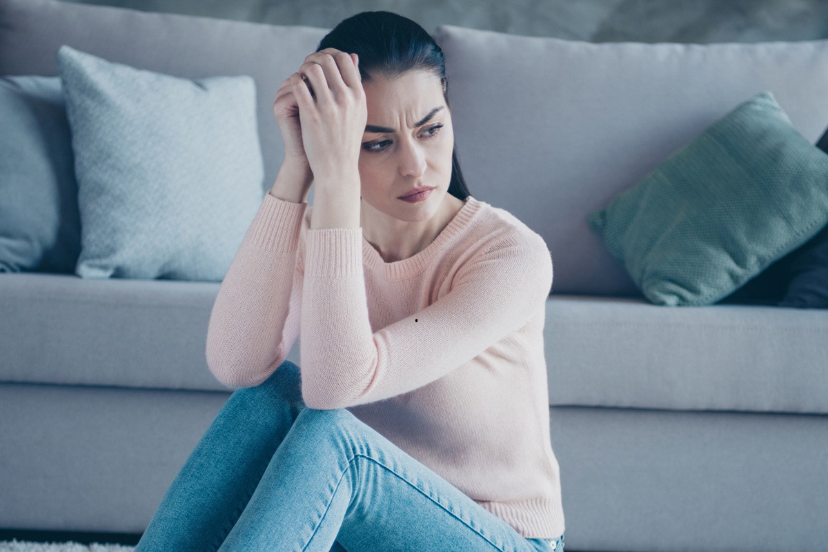 kobieta w wieku 30 plus w różowym sweterku siedzi na podłodze przy kanapie zmartwiona