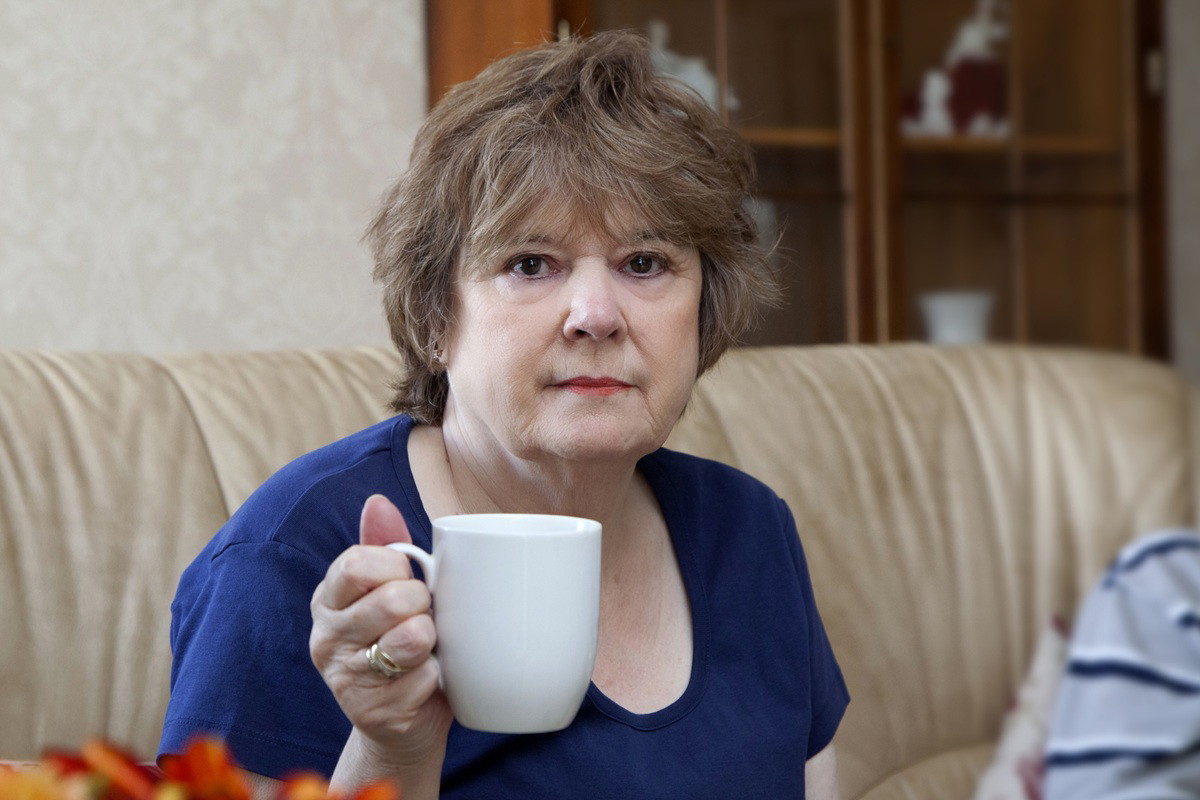 kobieta w wieku około 55 plus w niebieskiej koszulce siedzi na kanapie z kubkiem herbaty zamyślona