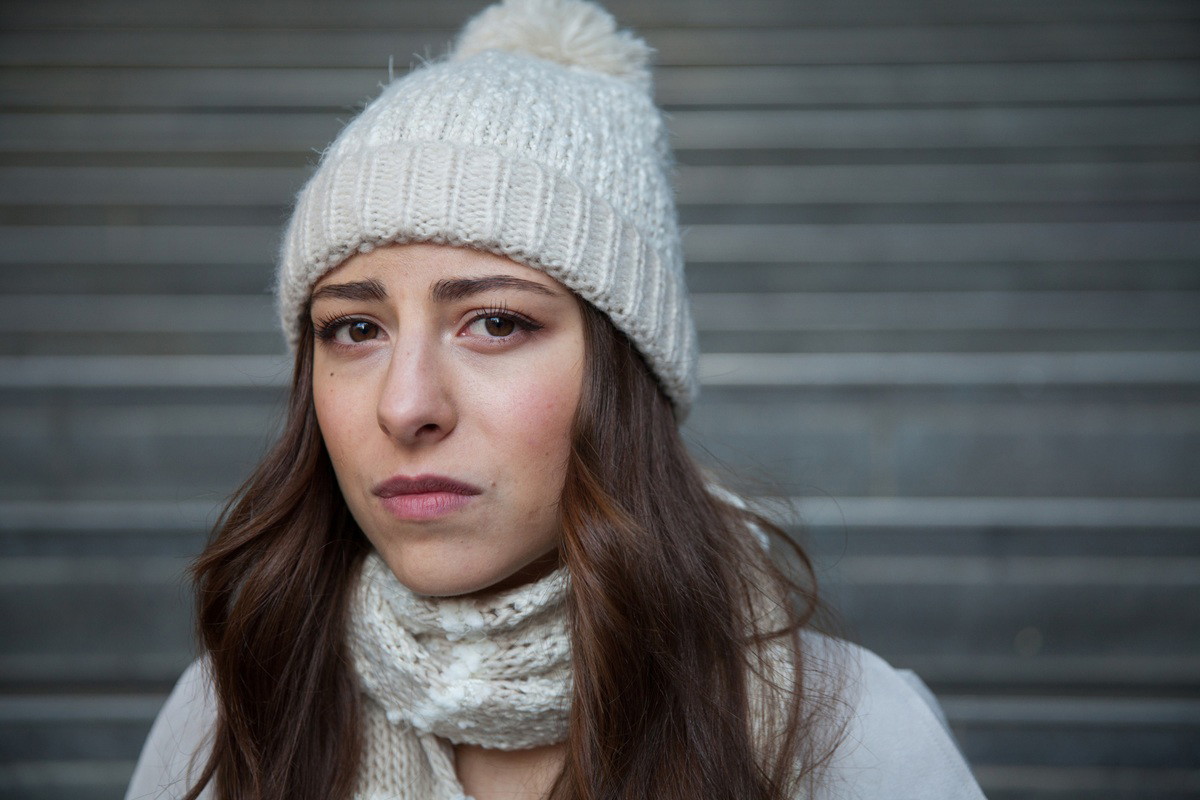 kobieta w wieku 30 plus w jasnej czapce z pomponem na tle metalowej ściany z niepewną miną