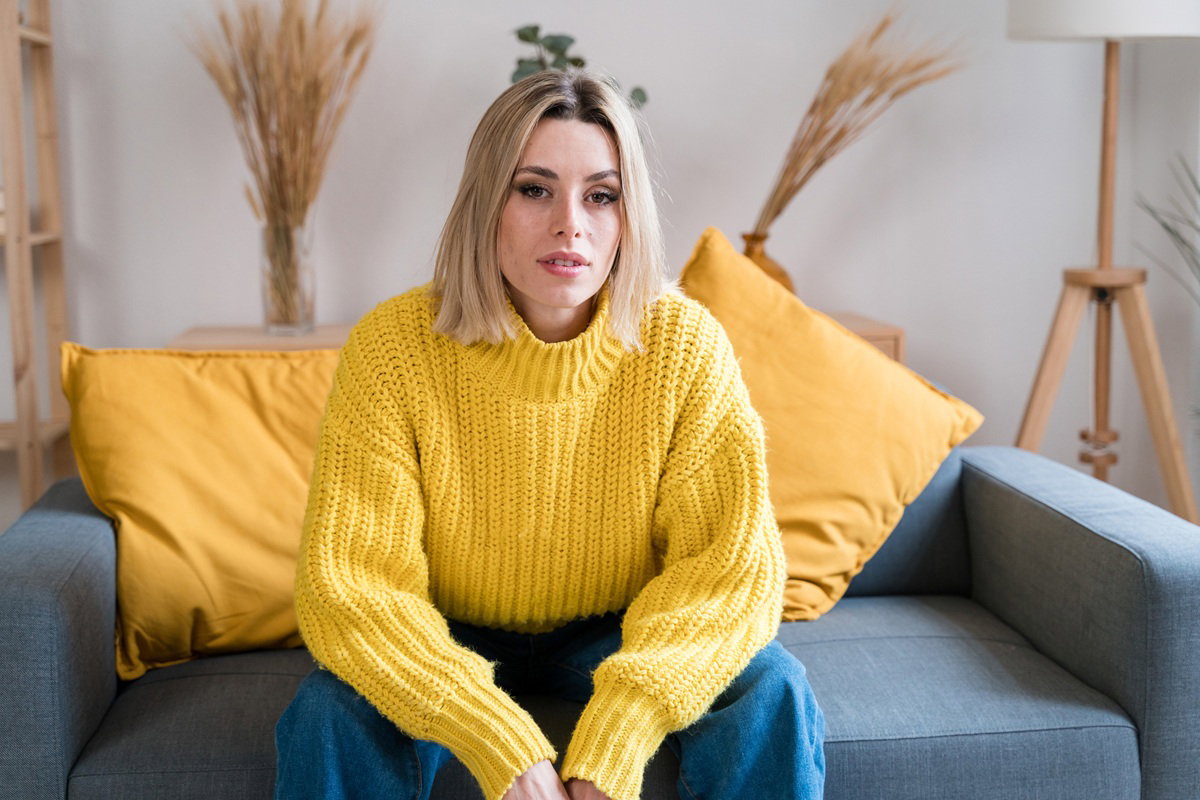 kobieta w wieku 30 plus blondynka w żółtym swetrze siedzi na kanapie zamyślona i wzruszona