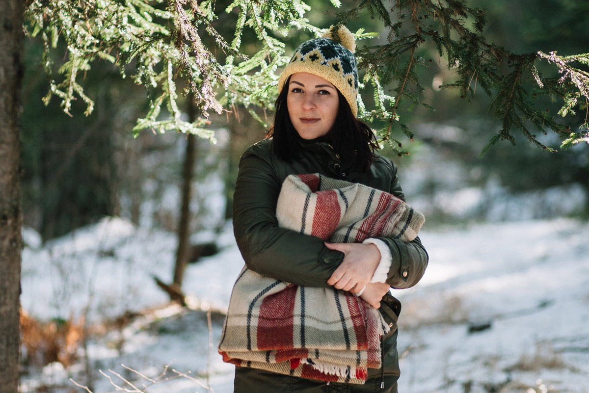 kobieta w wieku 30 plus w czapce z pomponem stoi na tle zimowego lasu z kocem pod pachą zamyślona