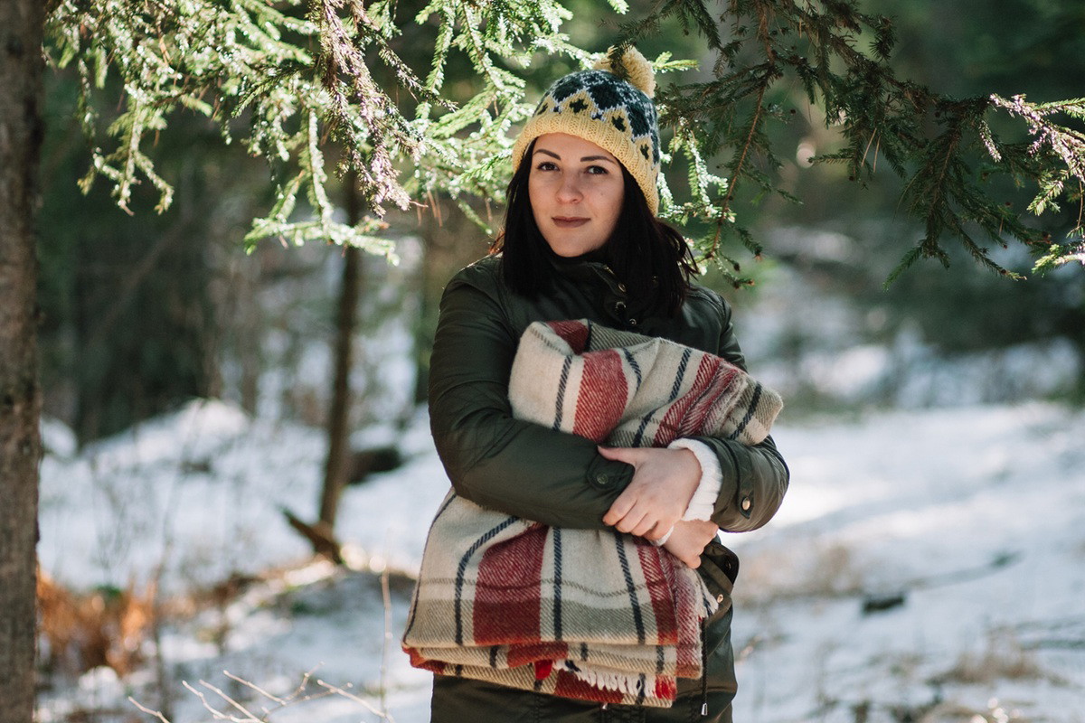 kobieta w wieku 30 plus w czapce z pomponem stoi na tle zimowego lasu z kocem pod pachą zamyślona