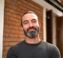 mężczyzna w wieku 37 lat z brodą i wąsami w ciemnym t-shircie stoi na tle domu i się uśmiecha
